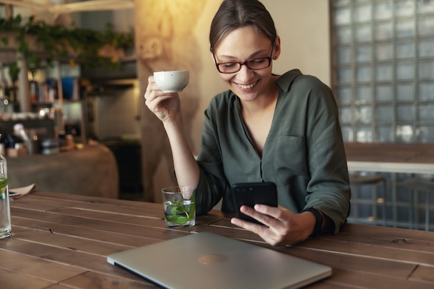 Femme à lunettes élégantes et veste noire, boire du café dans un restaurant et écrire du texte sur téléphone mobile