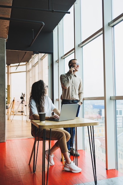 Photo une femme à lunettes est assise à une table et travaille sur un ordinateur portable un collègue masculin en arrière-plan parle au téléphone une journée de travail dans un bureau moderne