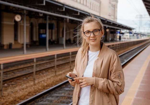 Femme avec des lunettes sur le quai près du train. Fille avec un téléphone portable à la gare près du train bleu. Voyage en train. Bydgoszcz.