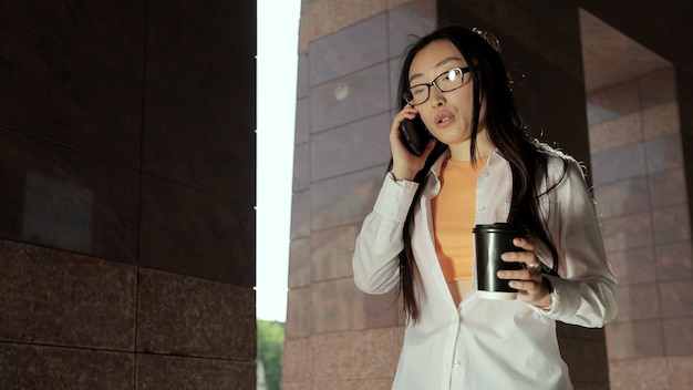 Une femme avec des lunettes s'approche de l'immeuble de bureaux et parle au téléphone, une jeune fille asiatique fait une pause tout en travaillant avec du café dans la rue