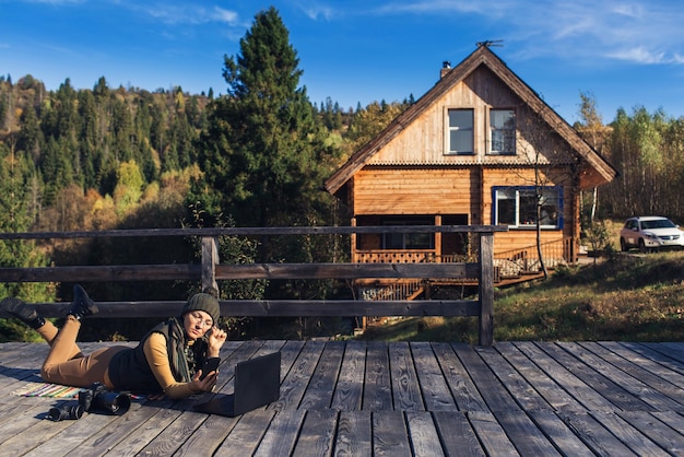 Une femme à lunettes se trouve sur un plaid en automne sur une terrasse en bois travaille sur un ordinateur portable et parle au téléphone