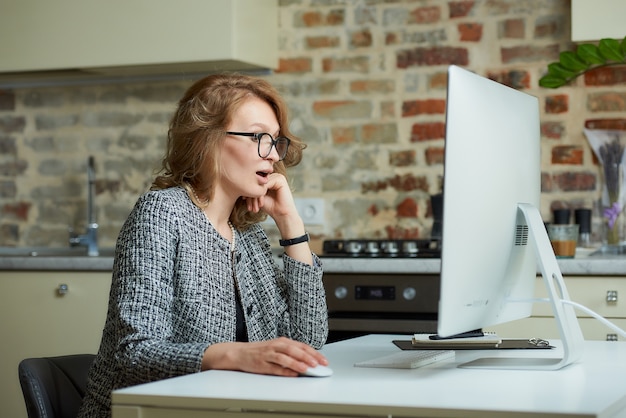Une femme à lunettes travaille à distance sur un ordinateur de bureau dans son studio. Une femme patron est surprise par des employés lors d'une vidéoconférence à domicile.