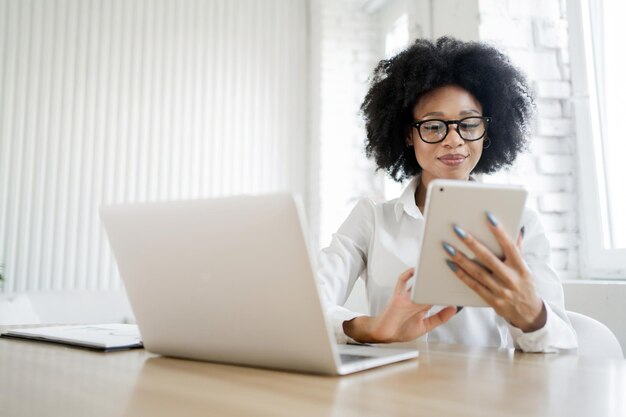 Une femme avec des lunettes travaille en ligne au bureau utilise une tablette et un ordinateur portable