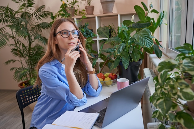 Femme à lunettes utilisant un téléphone tout en parlant pendant l'apprentissage en ligne à la maison