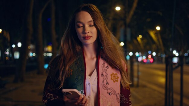 Une femme luxueuse allumant les lumières de la rue, la ville du soir, tenant un smartphone en gros plan.