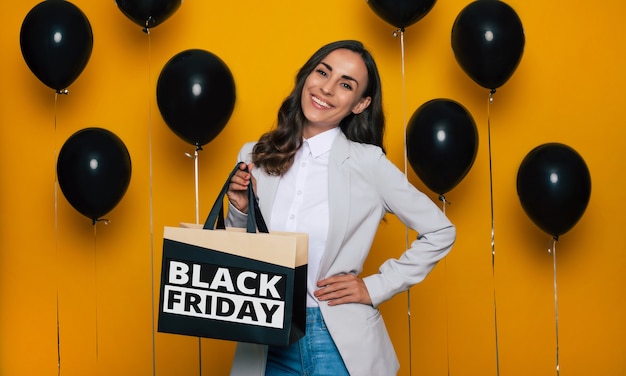 Une femme magnifique et heureuse à la mode avec un sac à provisions à la main pose dans une bonne humeur excitée à l'arrière-plan de nombreux ballons à l'hélium le vendredi noir