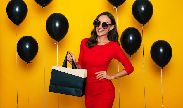 Une femme magnifique et heureuse à la mode avec un sac à provisions à la main pose dans une bonne humeur excitée à l'arrière-plan de nombreux ballons à l'hélium le vendredi noir