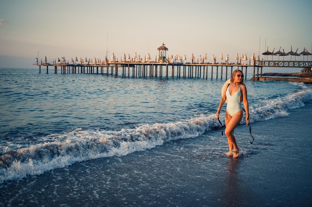 Une femme en maillot de bain et un chapeau à lunettes se promène le long de la plage au coucher du soleil Le concept de loisirs en mer Mise au point sélective