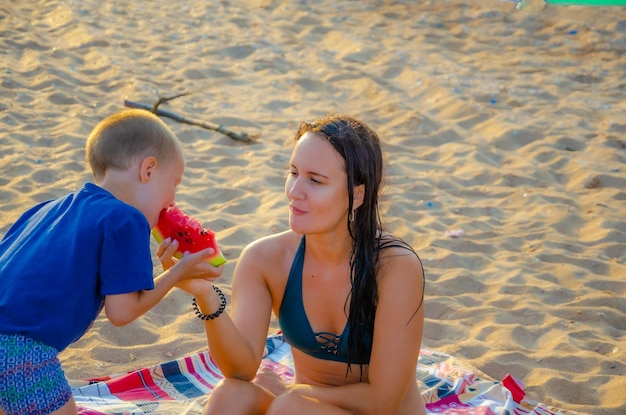 Une femme en maillot de bain sur la plage nourrit son fils une pastèque.