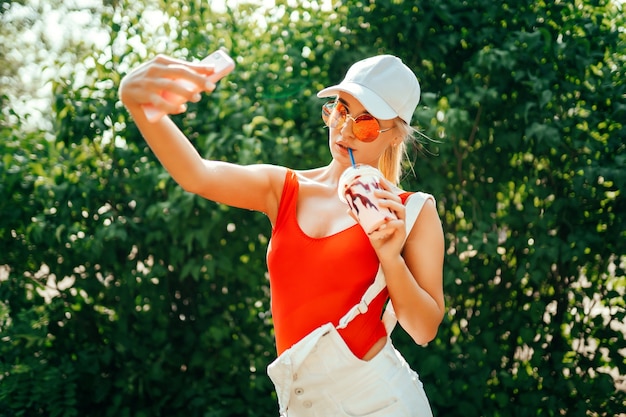 Femme en maillot de bain rouge et casquette blanche prenant un selfie et buvant un smoothie au parc