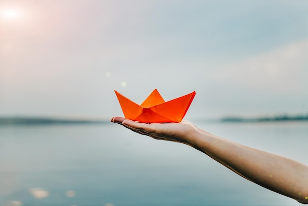 Femme main tenant un bateau en papier orange sur le fond de la rivière Une main d'une femme tenant un bateau en origami le soir près de l'eau