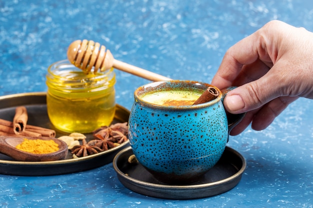 Femme main tenant la tasse de boisson ayurvédique traditionnelle lait de curcuma doré et assiette avec ses ingrédients sur bleu.