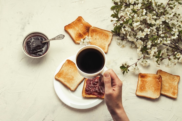 Femme main tenant une tasse de café, confiture, pain grillé, arbre de branches de printemps avec des fleurs. Concept de petit déjeuner. Mise à plat, vue de dessus