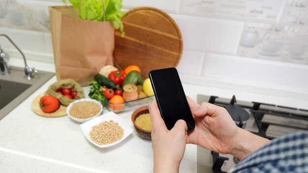 Femme main tenant un téléphone portable avec un écran vide sur une table de cuisine en arrière-plan avec des légumes d'épicerie