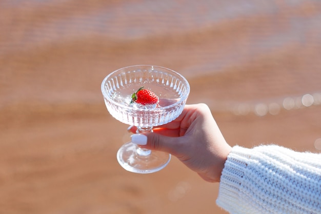 Femme main tenant un verre d'eau avec des fraises sur la mer dans le fond de sable