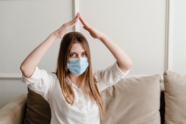 Femme à la maison sur un canapé portant un masque montrant le toit avec ses mains