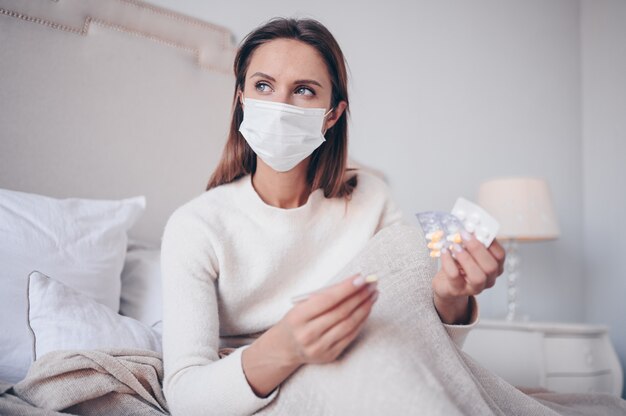 Femme malade dans un masque de protection du visage allongé dans son lit tenant un thermomètre et des pilules à la maison isolement de quarantaine.