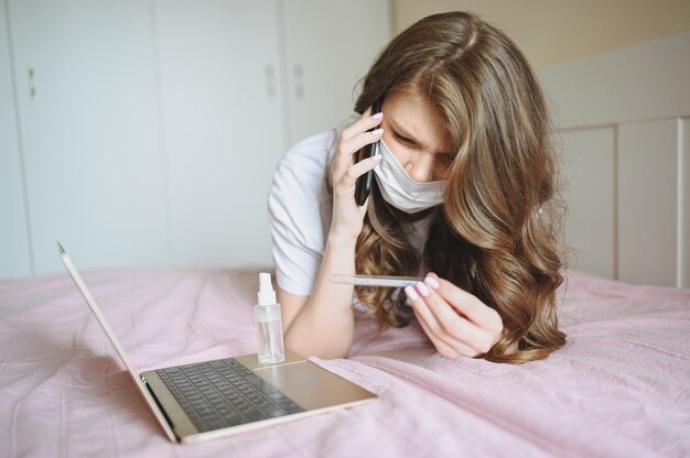 Femme malade en masque de protection du visage couché dans son lit avec un ordinateur portable et un désinfectant tenant un thermomètre