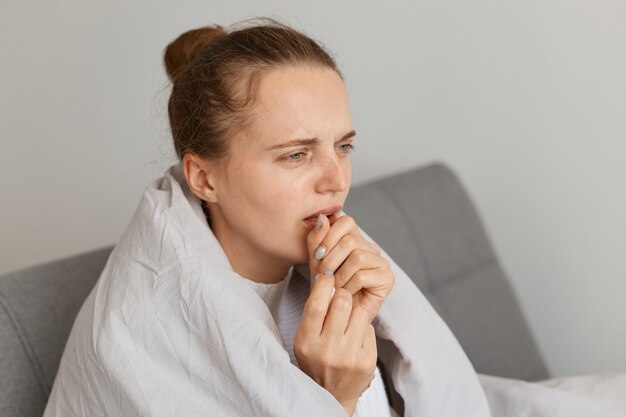 Femme malade en mauvaise santé avec un chignon assise sur un canapé enveloppé dans une couverture toussant, détournant les yeux avec une expression triste, tombe mal, symptômes de la grippe, posant à la maison.