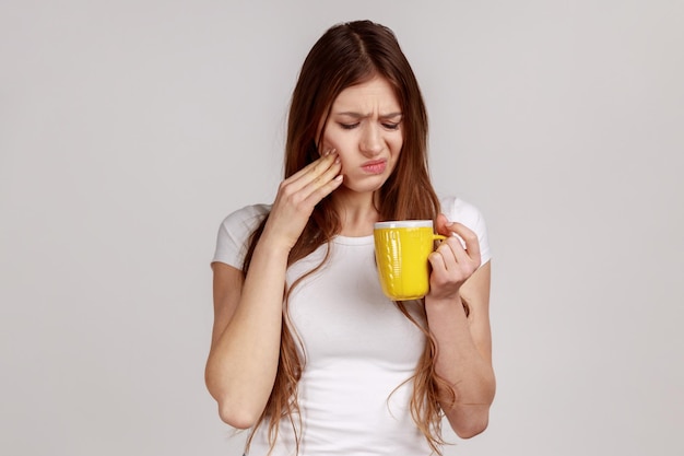 Femme malade en mauvaise santé touchant le menton souffrant de maux de dents, a des dents sensibles, ressent de la douleur après avoir bu de l'eau froide, portant un T-shirt blanc. Tourné en studio intérieur isolé sur fond gris.