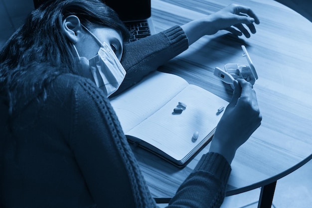 Femme malade avec des pilules et un thermomètre sur une table travaillant sur un ordinateur portable de la maison en quarantaine