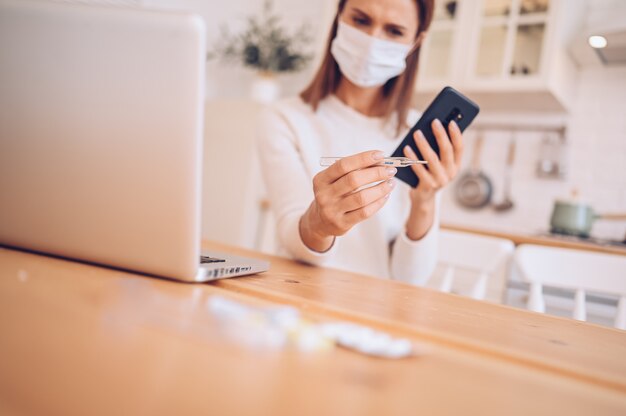 femme malade, porter, masque médical, parler, smartphone, et, travailler, a, ordinateur portable, dans, cuisine, maison, pendant, quarantaine, isolement, Covid-19, pandémie