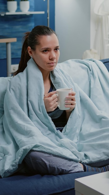Femme malade tenant une tasse de thé et assise dans une couverture. Personne ayant froid avec la maladie ayant des boissons et des médicaments, des bouteilles de pilules et des mouchoirs sur la table. Adulte malade