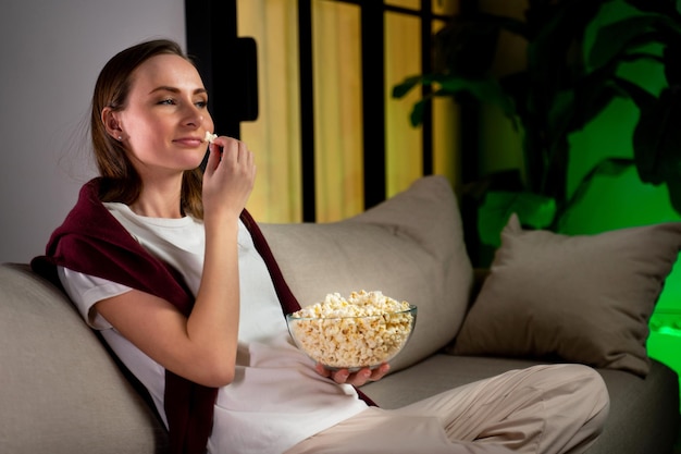 Une femme mange du pop-corn et regarde la télévision assise sur le canapé à la maison