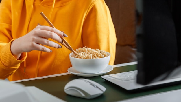 Femme mange des nouilles avec des baguettes près de la souris et du clavier pc ordinateur portable dans le bureau à domicile