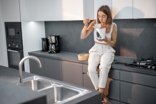 Une femme mange des plats à emporter tout en étant assise sur la cuisine moderne