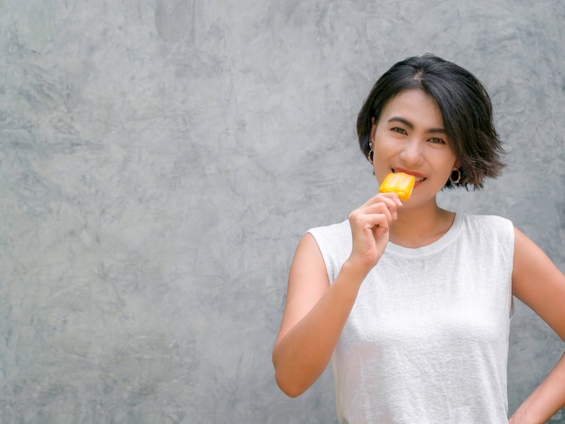 Femme mangeant des sucettes glacées. Heureuse belle femme asiatique cheveux courts portant une chemise sans manches blanche décontractée tenant un popsicle jaune sur fond de mur de béton gris avec espace de copie, en été.