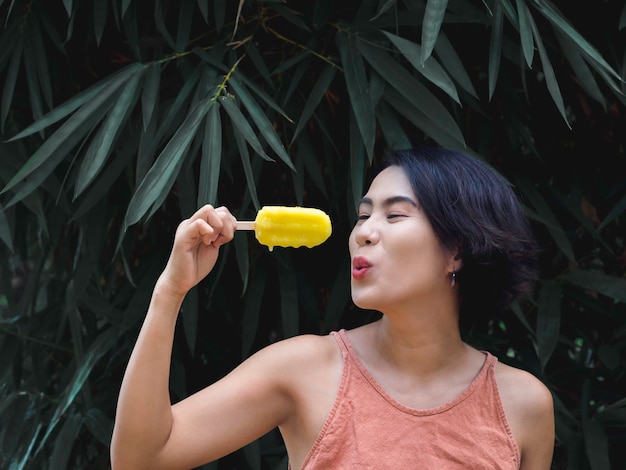 Femme mangeant des sucettes glacées. Heureuse belle femme asiatique portant un débardeur rose décontracté tenant un popsicle jaune sur fond de feuilles de palmier tropical vert, en plein air. Femme souriante appréciant la sucette glacée en été.