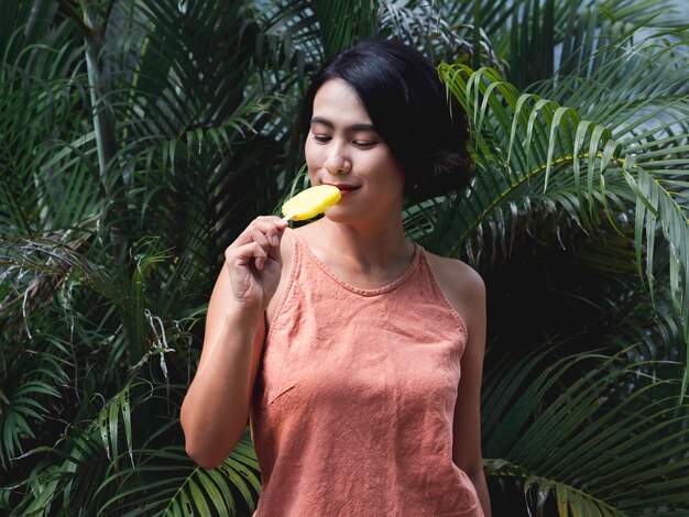 Femme mangeant des sucettes glacées. Heureuse belle femme asiatique portant un débardeur rose décontracté tenant un popsicle jaune sur fond de feuilles de palmier tropical vert, en plein air. Femme souriante appréciant la sucette glacée en été.