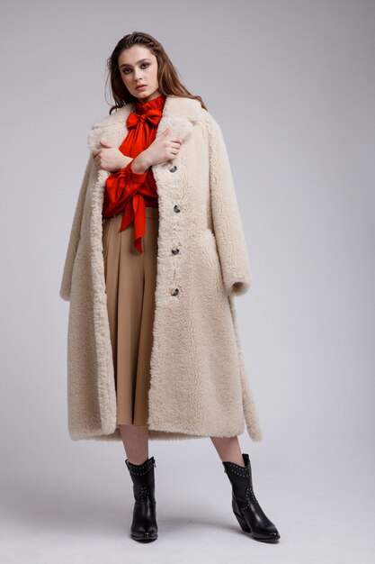 Femme en manteau beige jupe longue chemisier arc rouge bottes sur fond blanc cheveux bruns Studio Shot