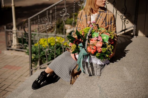 Femme en manteau à carreaux assis sur des escaliers en béton avec un panier de fleurs en osier