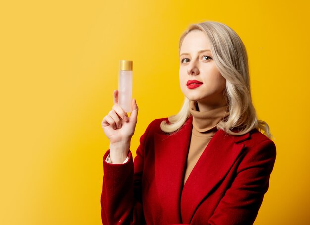 Femme en manteau rouge avec bouteille de lotion sur mur jaune