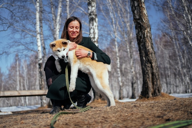 Une femme en manteau sombre embrasse un chiot akita inu avec une longue laisse verte dans un parc en automne