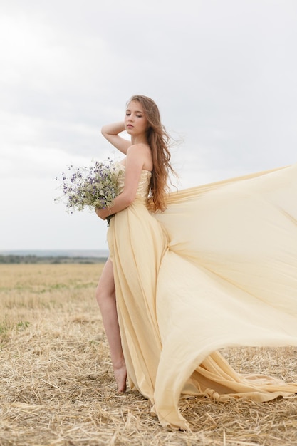 Femme marchant dans le champ d'herbe séchée dorée. Beauté naturelle du portrait. Belle fille dans un champ de blé. Jeune femme en robe beige tenant un bouquet de fleurs sauvages.
