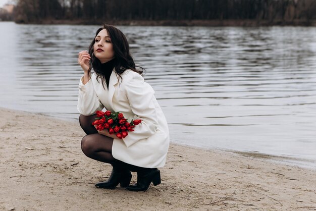 Femme marchant sur le rivage de la plage dans un temps tempéré et venteux avec un bouquet de fleurs de tulipes rouges