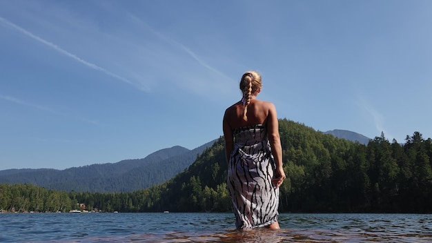 Femme marche sur l'eau sur la jetée en lunettes de soleil et un châle en soie bohème Fille se repose sur un quai sous-marin en bois d'inondation Le trottoir est recouvert d'eau dans le lac En arrière-plan se trouvent la montagne et une forêt