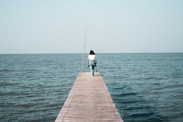 Femme marche seule sur un pont en bois dans la mer avec un ciel bleu.