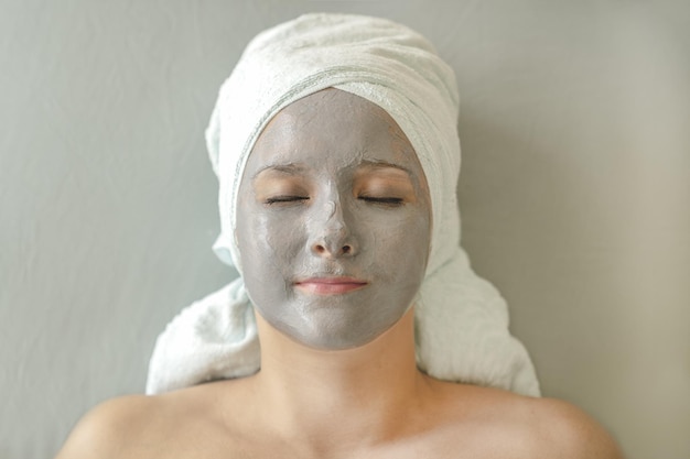 Femme avec un masque d'argile sur le visage et une serviette sur la tête se trouve et bénéficie d'un traitement spa pour les soins de la peau du visage