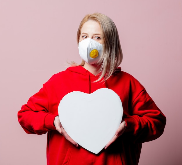 Femme en masque facial anti-poussière FFP2 tenir la bannière en forme de coeur sur fond rose