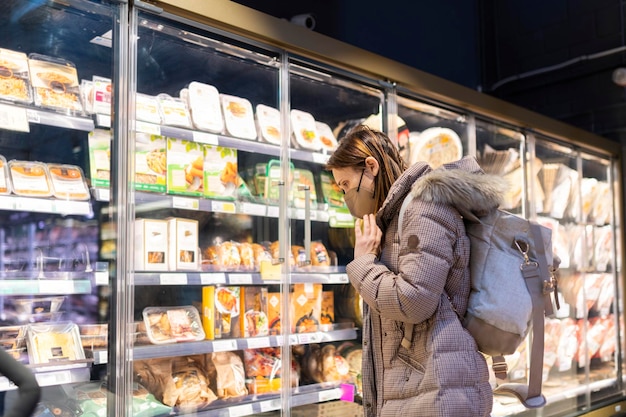 Femme avec un masque facial faisant les courses au supermarché dans la zone des plats précuits et emballés aliments transformés