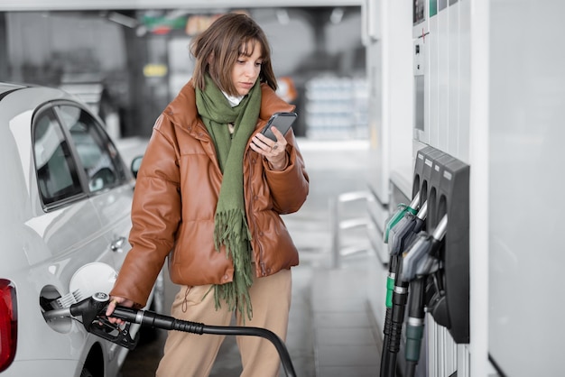 Femme en masque facial faisant le plein d'une voiture avec de l'essence à l'aide d'un téléphone intelligent