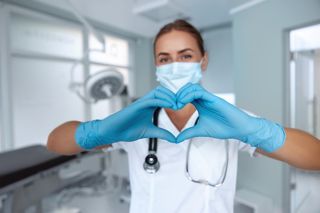 Une femme avec un masque médical et des mains dans un gant en latex montre le symbole du coeur