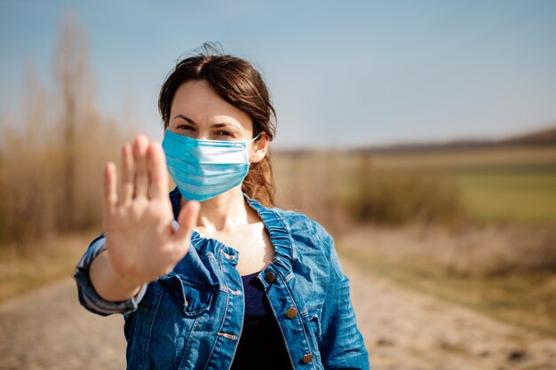 Femme en masque médical stérile protecteur sur son visage regardant la caméra à l'extérieur. Panneau d'arrêt de la main. Concept de coronavirus pandémique.