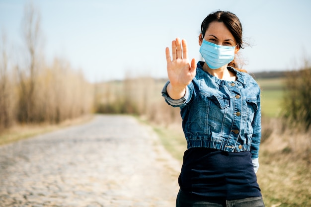 Femme en masque médical stérile protecteur sur son visage regardant la caméra à l'extérieur. Panneau d'arrêt de la main. Concept de coronavirus pandémique.