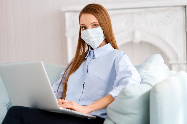 Femme en masque de protection médical travaillant sur un ordinateur portable en ligne de la maison assise sur le canapé