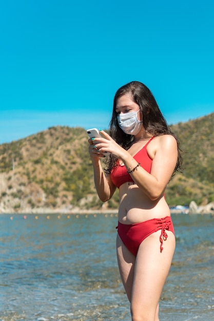 Femme avec masque de protection et téléphone en maillot de bain sur la plage